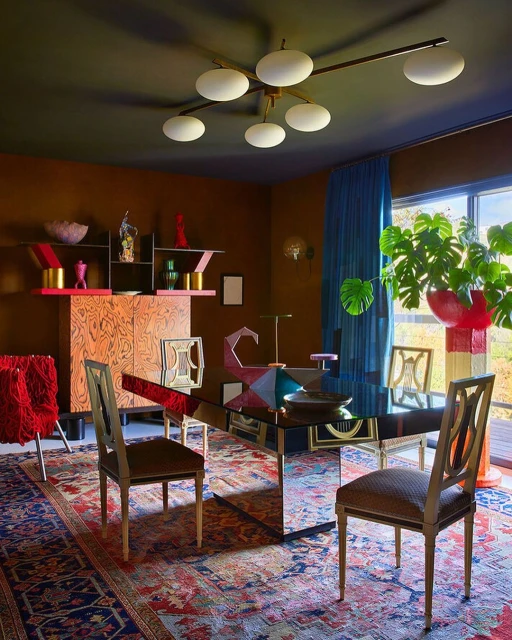 Eclectic dining room features a glossy black dining table, mixed chairs, geometric cabinet, blue curtain, and large potted plant.