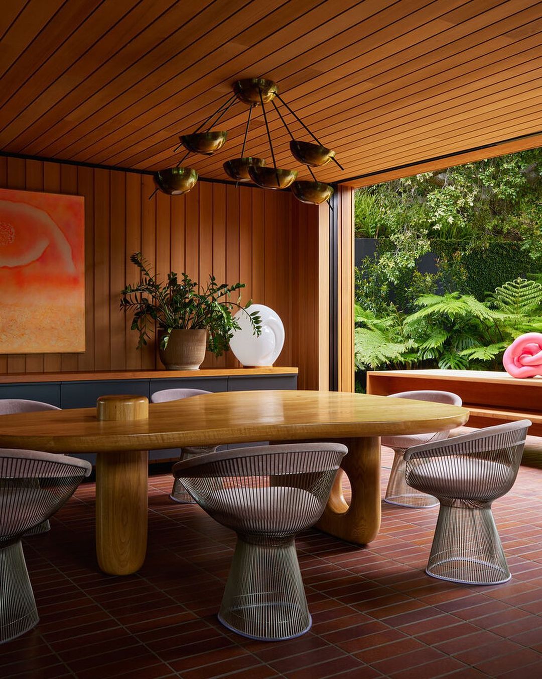 Indoor dining room featuring a wooden table and chairs with a view of a lush green garden. Warm lighting creates a cozy atmosphere.