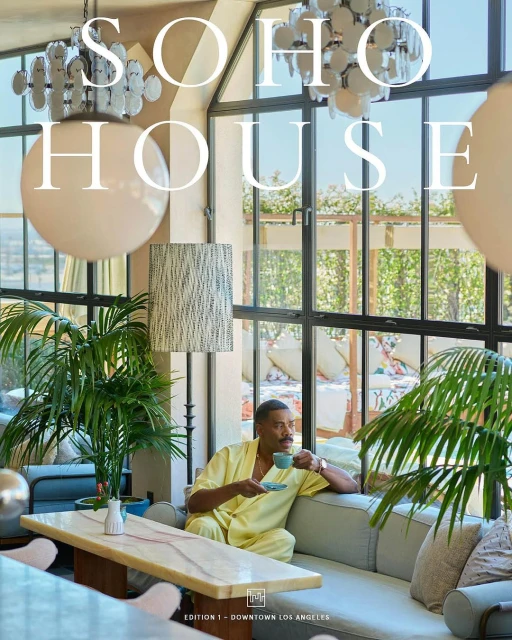 Adult male in a yellow robe seated on a sofa, holding a teacup in a bright indoor lounge setting with natural light and potted plants.