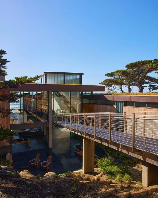 Exterior view of a modern, multi-level house with a connecting walkway, surrounded by trees and plants on a sunny day.