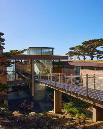 Exterior view of a modern, multi-level house with a connecting walkway, surrounded by trees and plants on a sunny day.