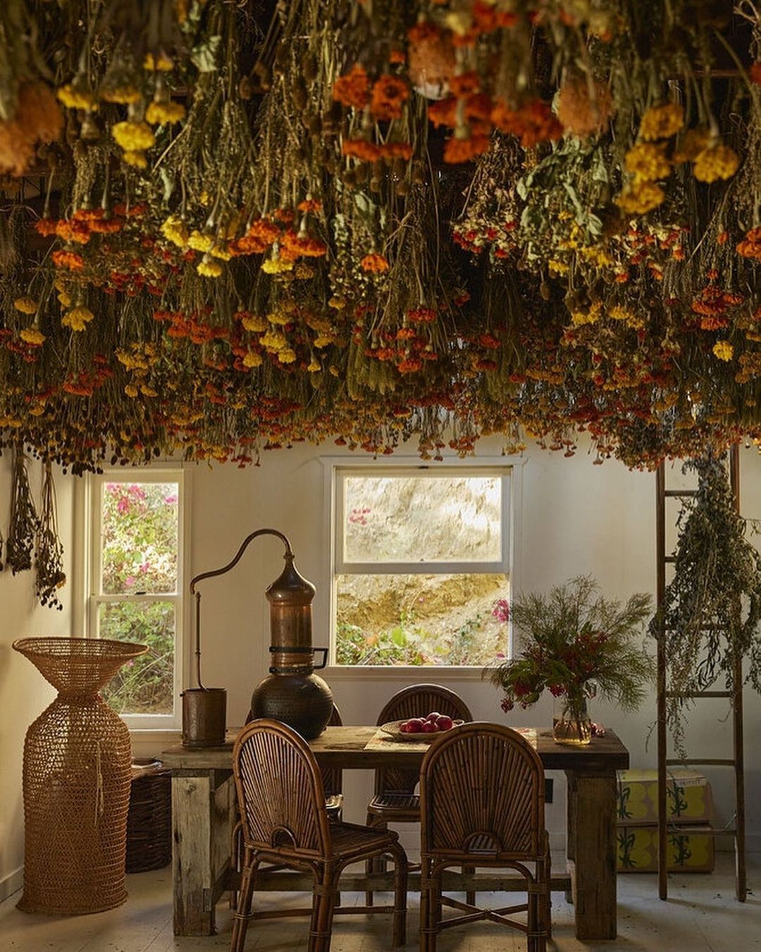 Dried flowers hang above a rustic wood table in a sunlit room, creating an enchanting and natural indoor setting with floral arrangements.