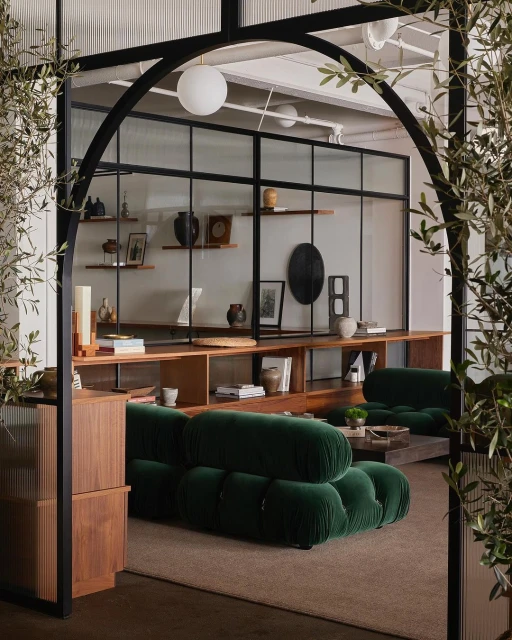 Living room features two green velvet sofas, wooden shelving, decor items, glass partition with black frame, and earth-tone carpet flooring.