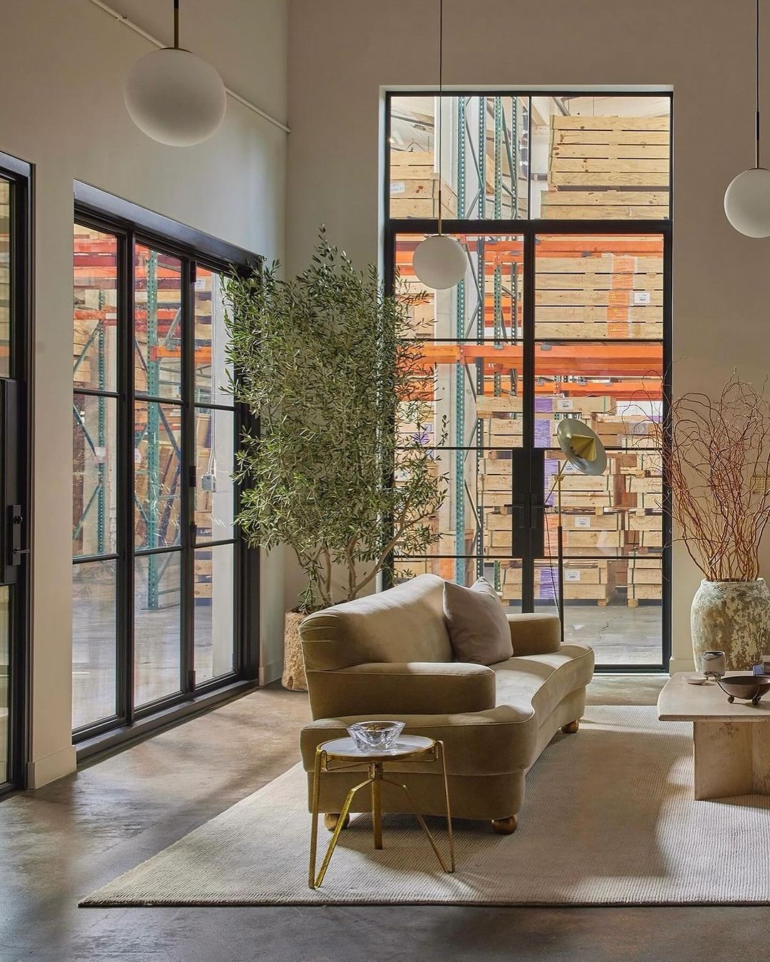 Living room interior with a beige sofa, a marble side table, and large windows overlooking a warehouse storage area.
