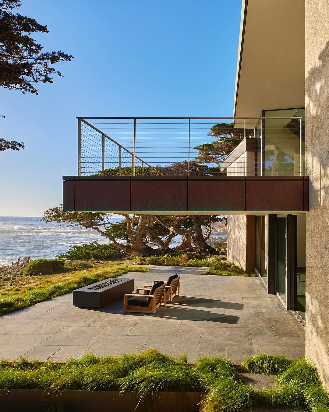 Exterior view of modern home with patio overlooking the ocean. Patio features outdoor furniture, a fire pit, and drought-tolerant greenery.