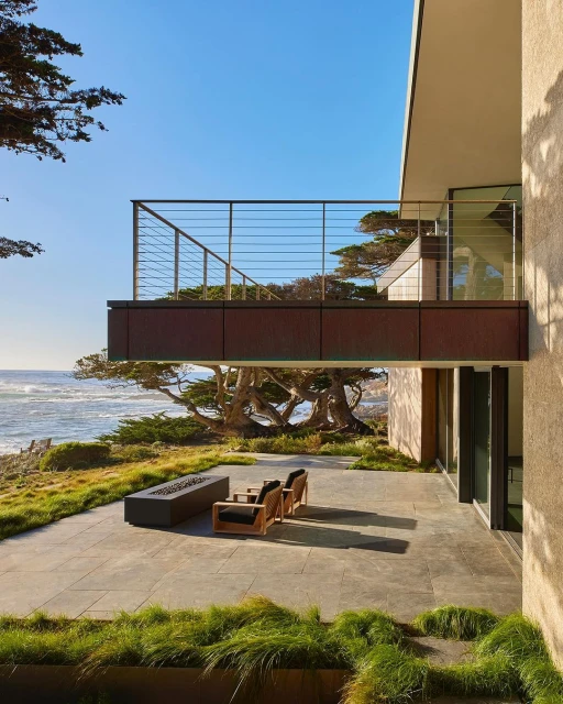 Exterior view of modern home with patio overlooking the ocean. Patio features outdoor furniture, a fire pit, and drought-tolerant greenery.