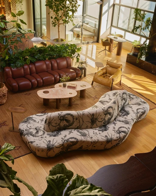 Interior view of a brightly lit living room featuring brown leather sofa, patterned chaise lounge, oval coffee tables, and houseplants.