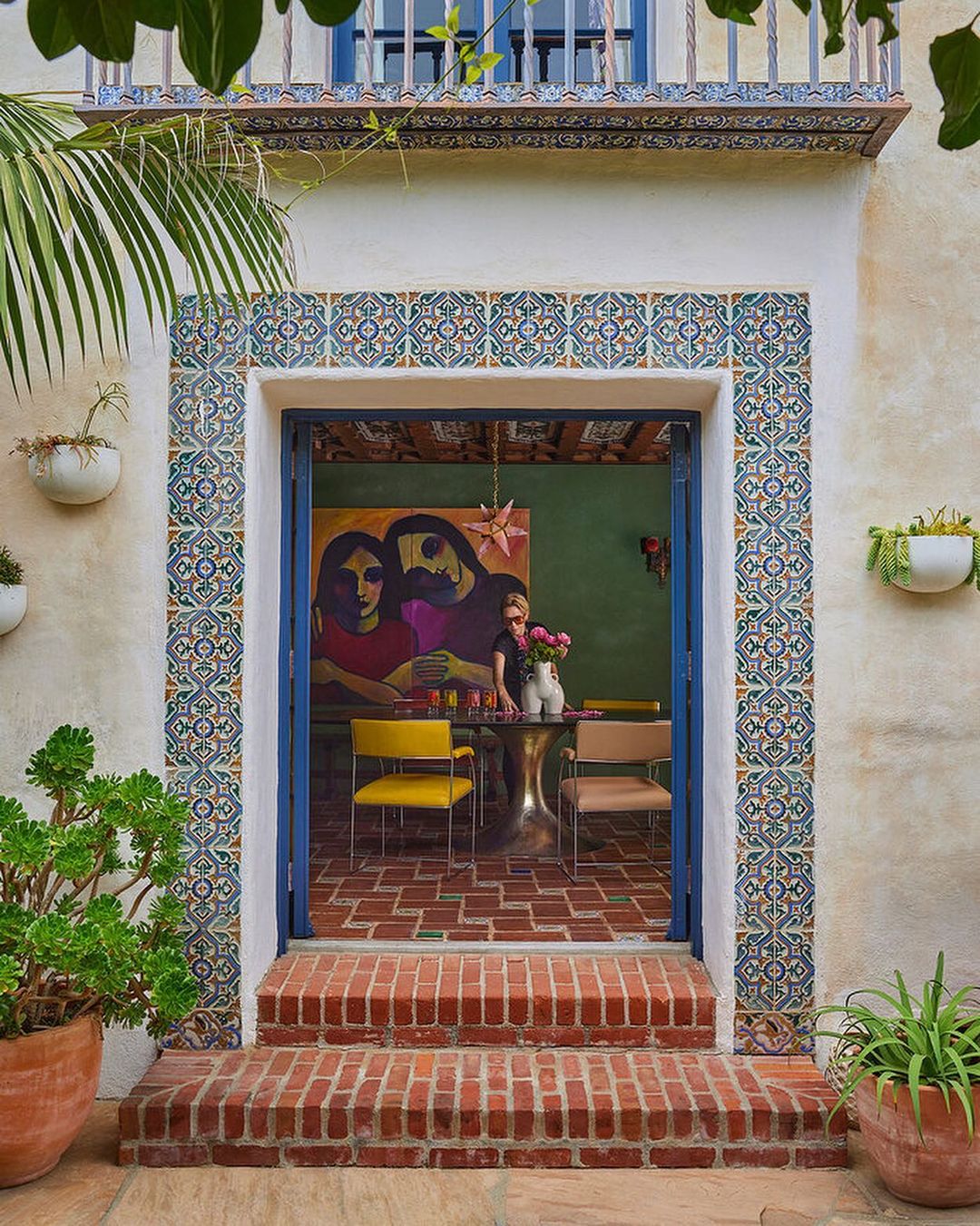 View through a doorway into an interior featuring a woman at a table, framed by tile-trimmed walls and potted plants.