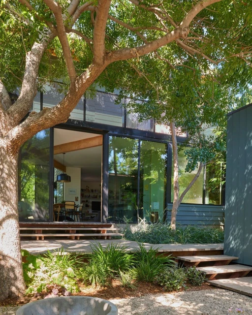 Exterior view of a modern house with large glass doors and windows, surrounded by trees and greenery in a natural setting.