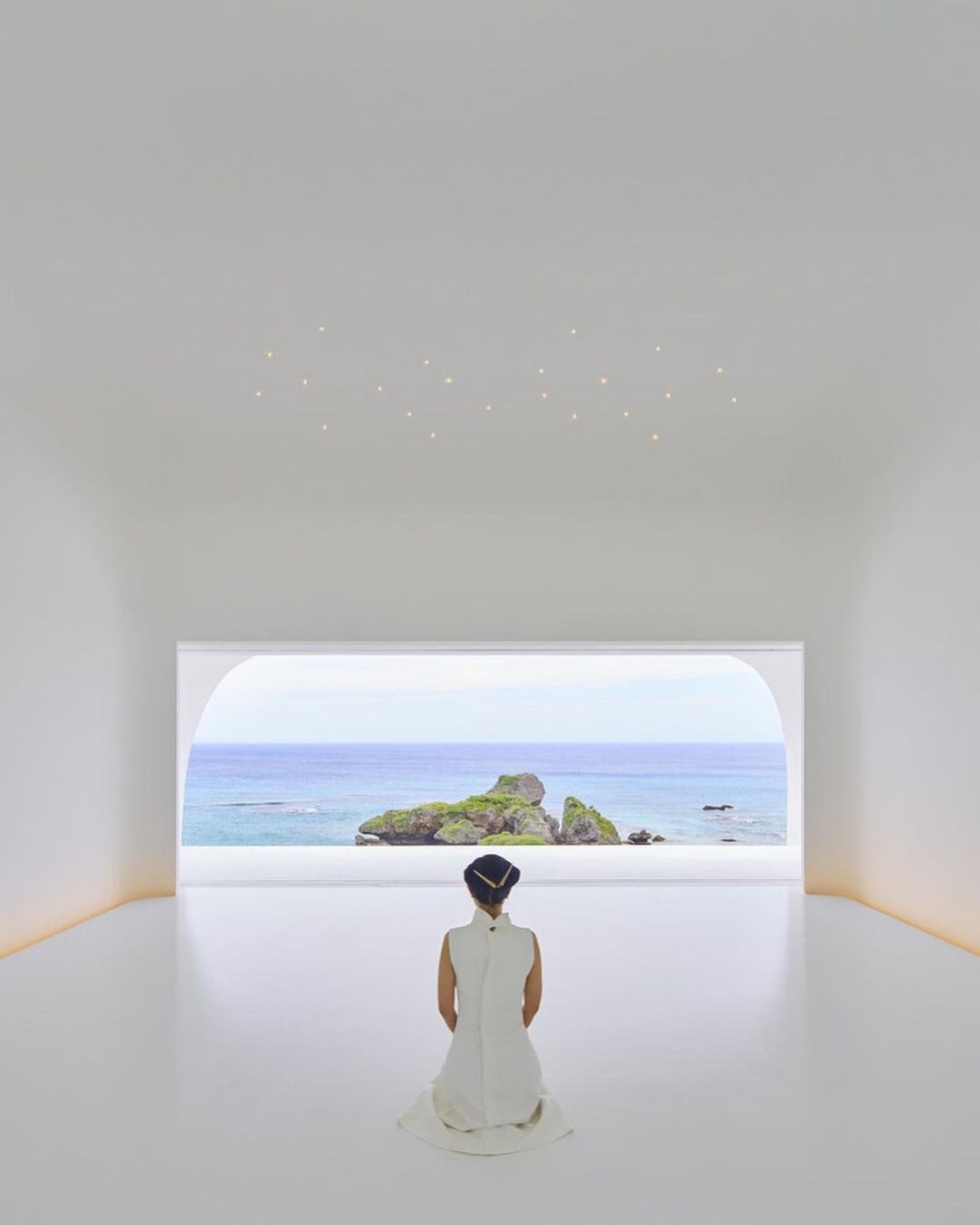 Woman in white dress sits in minimalist white room facing ocean view with island. Serene scene with clean lines and open space.