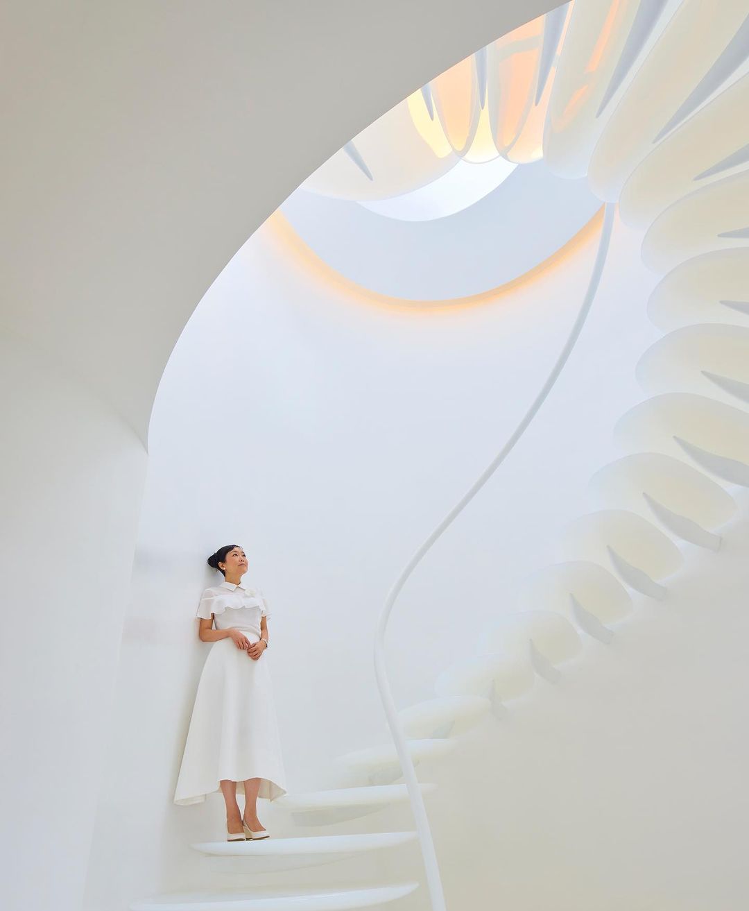 Adult female in white dress and shoes poses on a spiraling white staircase in a bright indoor setting with natural light overhead.
