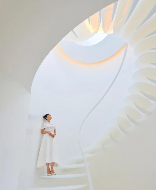 Adult female in white dress and shoes poses on a spiraling white staircase in a bright indoor setting with natural light overhead.