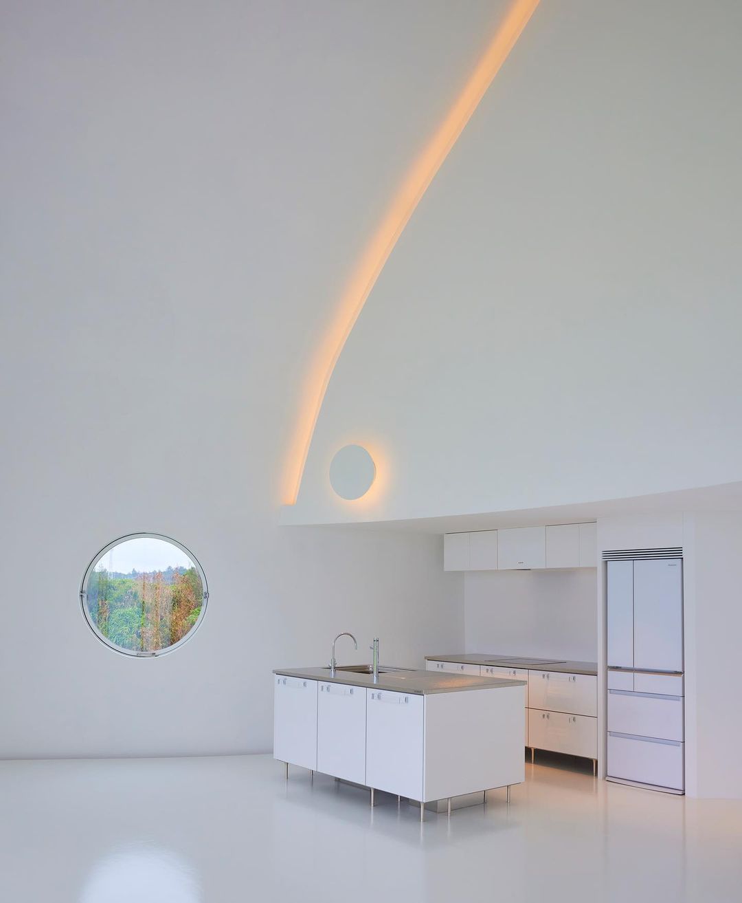 Modern white kitchen featuring island, cabinetry, stainless steel sink, round window with nature view, and indirect linear lighting.