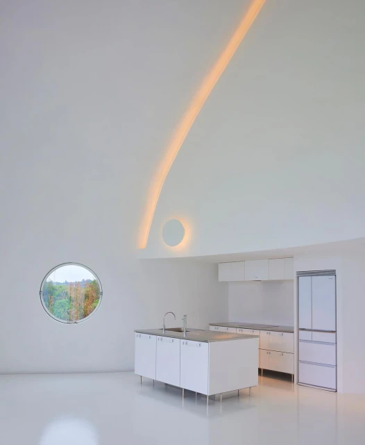 Modern white kitchen featuring island, cabinetry, stainless steel sink, round window with nature view, and indirect linear lighting.