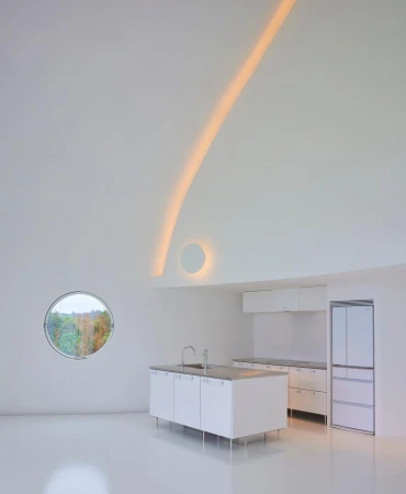 Modern white kitchen featuring island, cabinetry, stainless steel sink, round window with nature view, and indirect linear lighting.