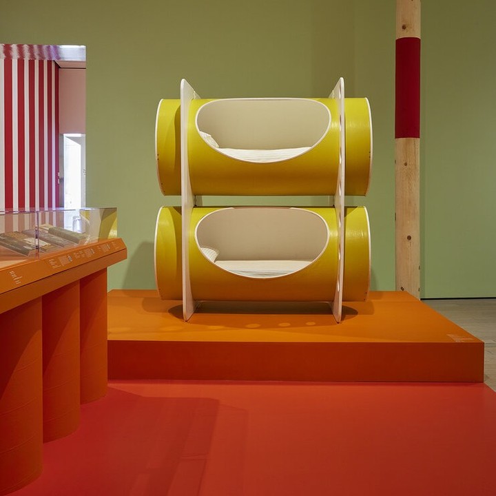 Modern art installation featuring yellow cylindrical forms stacked on an orange platform inside a gallery setting with colorful backdrop.