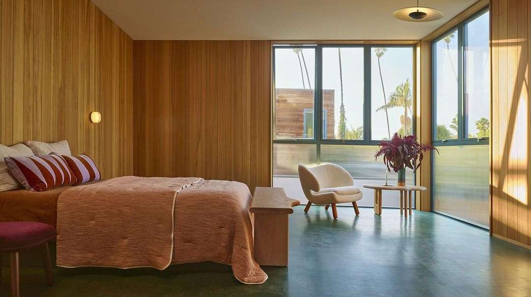 Bedroom interior featuring a bed with a patterned quilt, a modern chair, and sunlight streaming through large windows, creating a cozy scene.