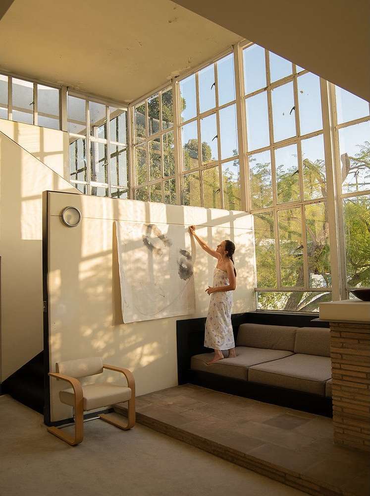 Woman stands on a sofa in a bright, modern interior, hanging a canvas with abstract designs on a wall with large windows.