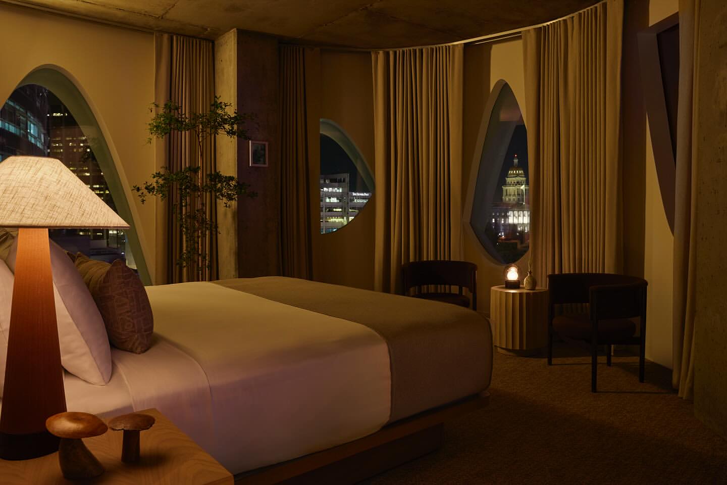 Cozy hotel room interior featuring a king-size bed, unique windows showcasing city lights at night, and decorative mushroom-shaped objects.