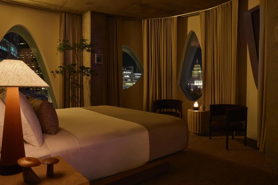 Cozy hotel room interior featuring a king-size bed, unique windows showcasing city lights at night, and decorative mushroom-shaped objects.