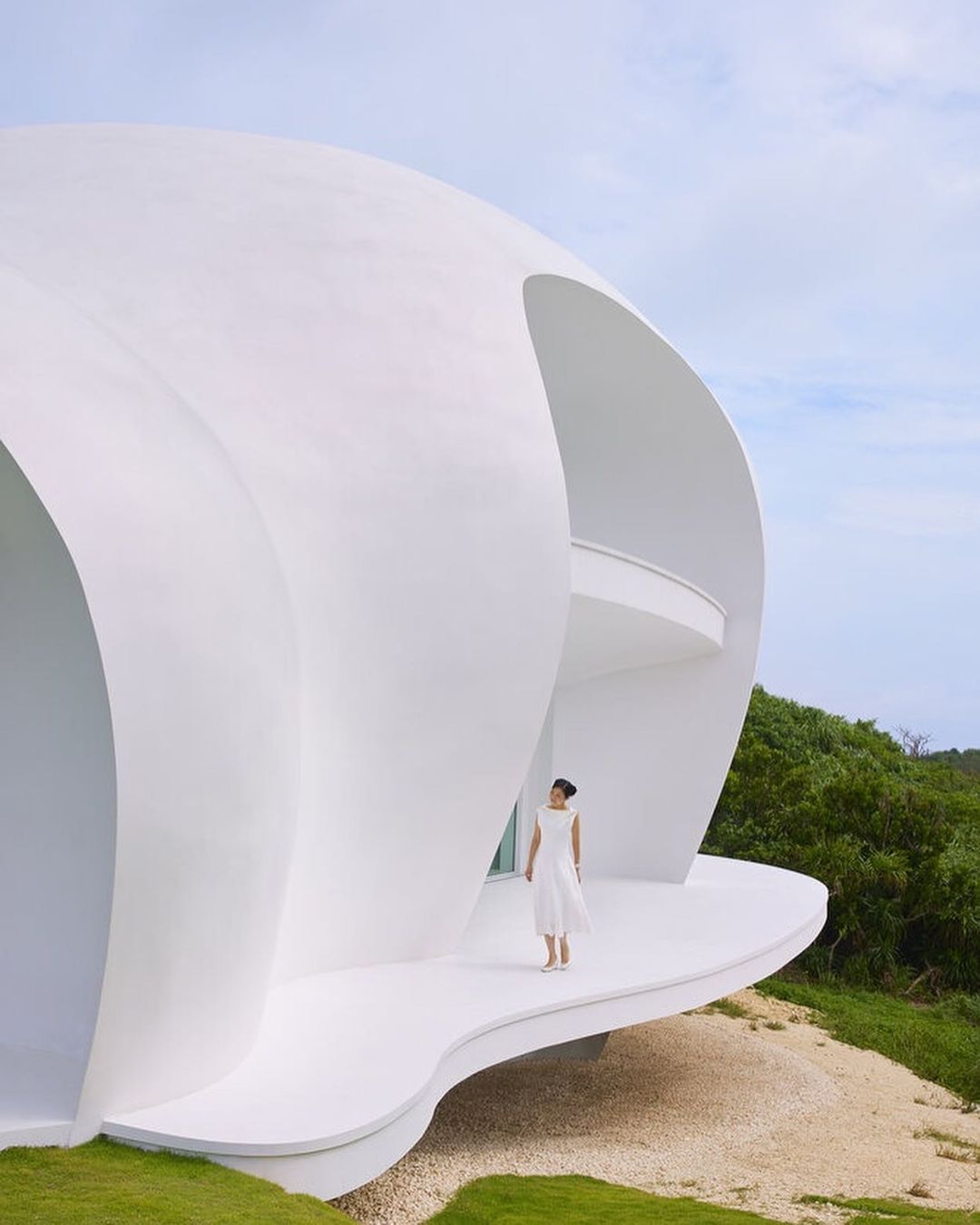 Adult woman in white dress stands on the white curved balcony of an architectural white shell-shaped modern house in natural light.
