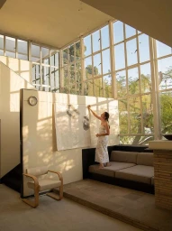 Woman in white dress hanging artwork on a wall in a bright modern living room with large grid windows by Yoshihiro Makino