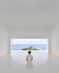 A person in a white dress kneeling in a minimalist white room with a large window looking out to the ocean, photographed by Yoshihiro Makino.