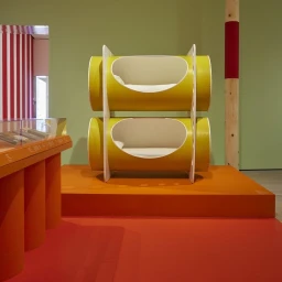 Yellow and white modular bunk bed sculpture displayed on an orange platform inside a gallery exhibit.