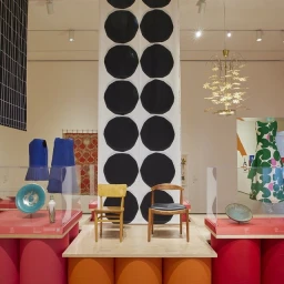 Exhibition interior featuring a central banner with large black circles, two wooden chairs, and various garments and ceramics on display
