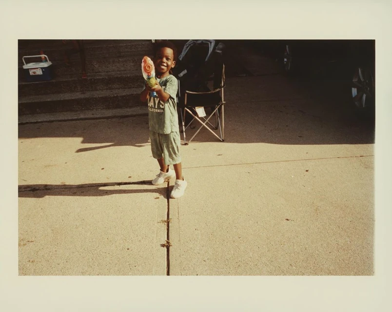 Young boy stands on driveway holding a water gun. Wearing shorts, t-shirt, and sneakers, he smiles on a sunny day outside.