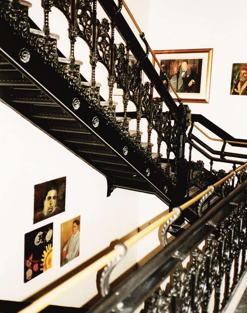 Black ornate staircase with wooden handrail and vintage portraits hanging on a white wall in the background.