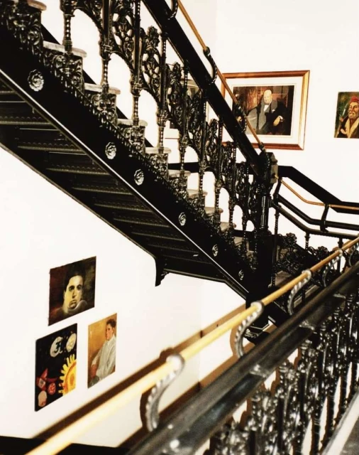 Black ornate staircase with wooden handrail and vintage portraits hanging on a white wall in the background.