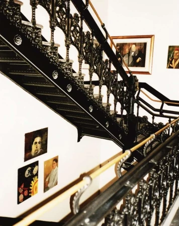 Black ornate staircase with wooden handrail and vintage portraits hanging on a white wall in the background.
