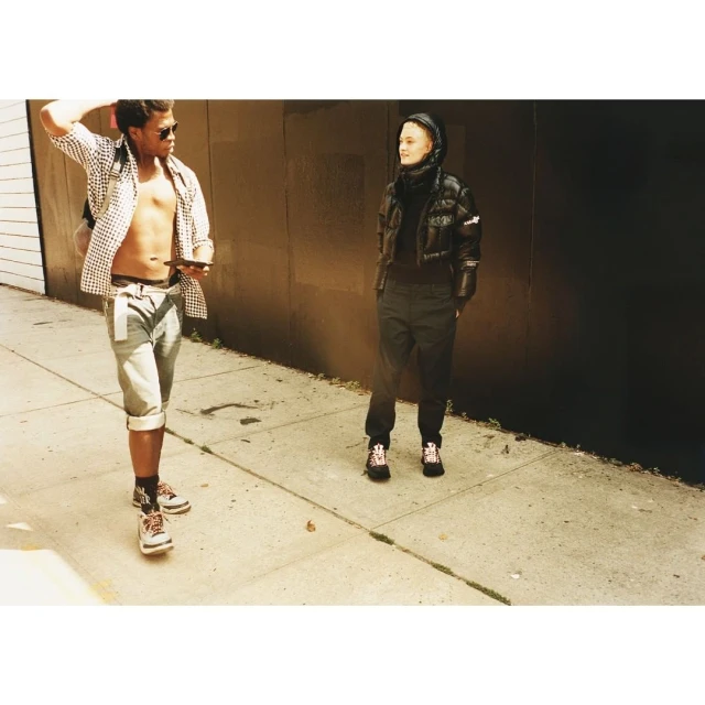 Two people in casual fashion stand outside on a city sidewalk with a dark wall; one wears a puff jacket, the other an unbuttoned checkered shirt.
