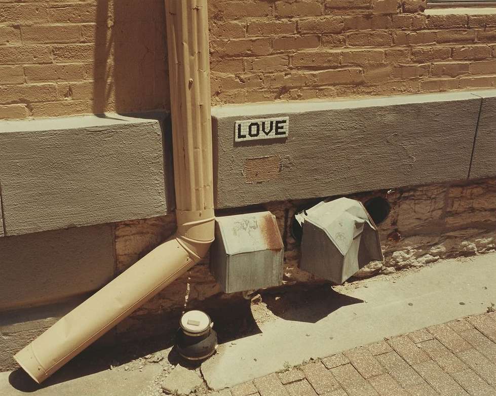Urban scene shows a brick building with a drainage pipe, a small water meter, and graffiti art of the word 'LOVE' visible on the wall.