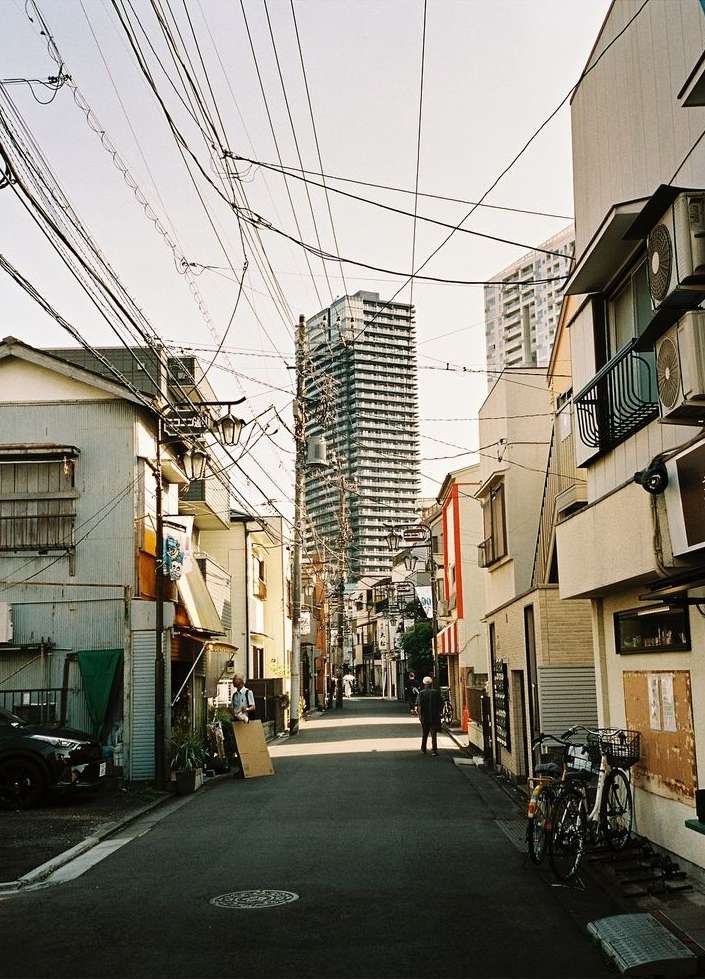 Urban scene showcases a narrow city street lined with buildings, power lines overhead, and two people walking in the distance.