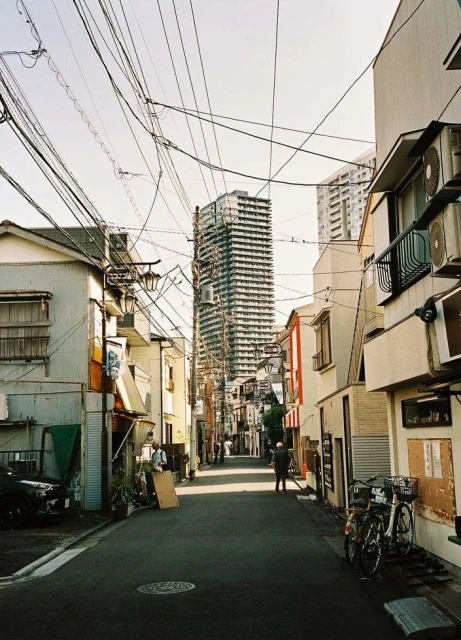 Urban scene showcases a narrow city street lined with buildings, power lines overhead, and two people walking in the distance.