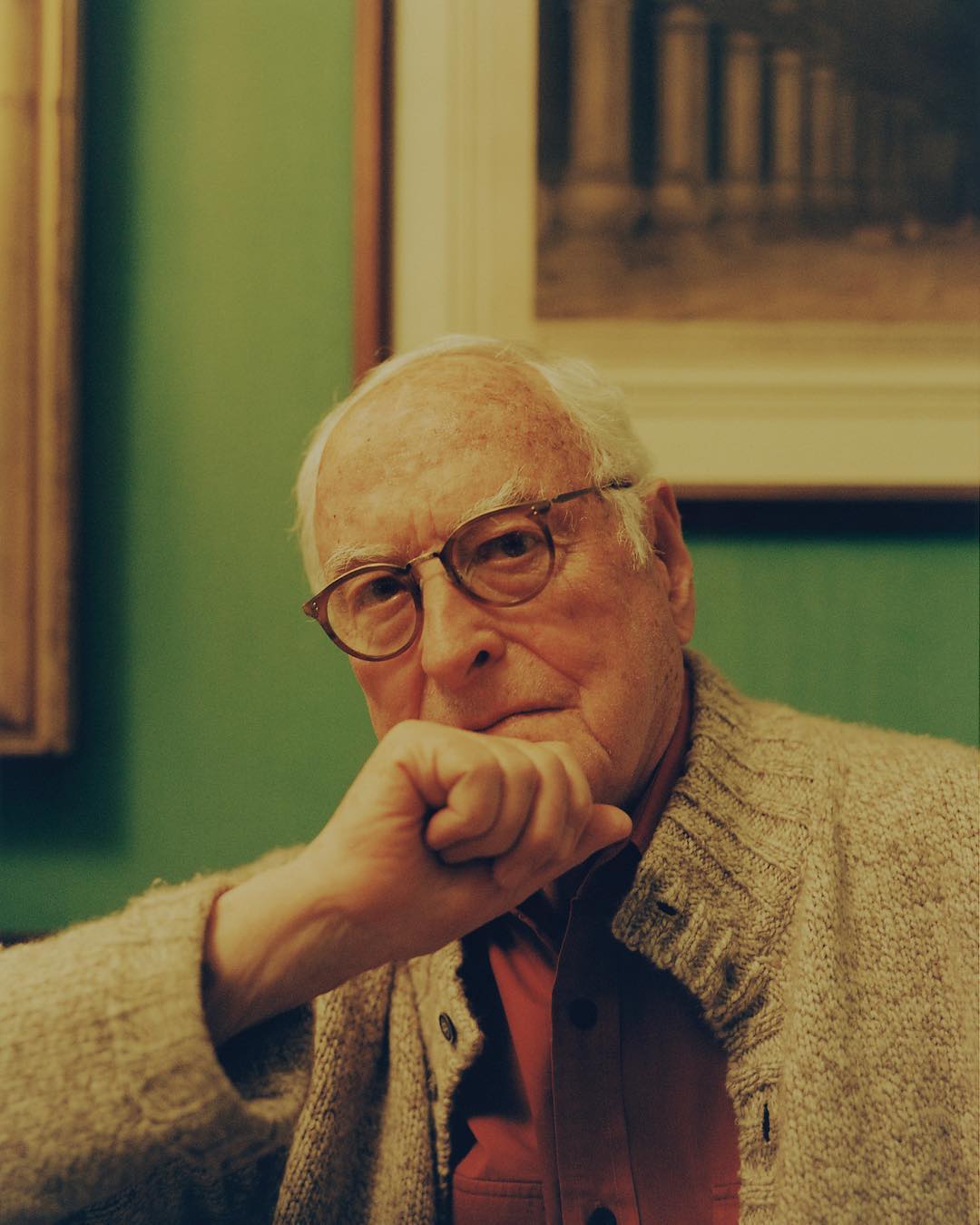 Portrait of an elderly man with glasses, wearing a cardigan, sitting indoors with artwork visible in the background on green walls.