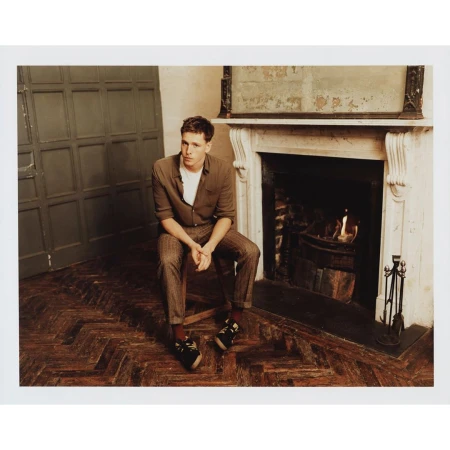A young man seated on a chair in a room with a herringbone wooden floor and an ornate fireplace mantel.