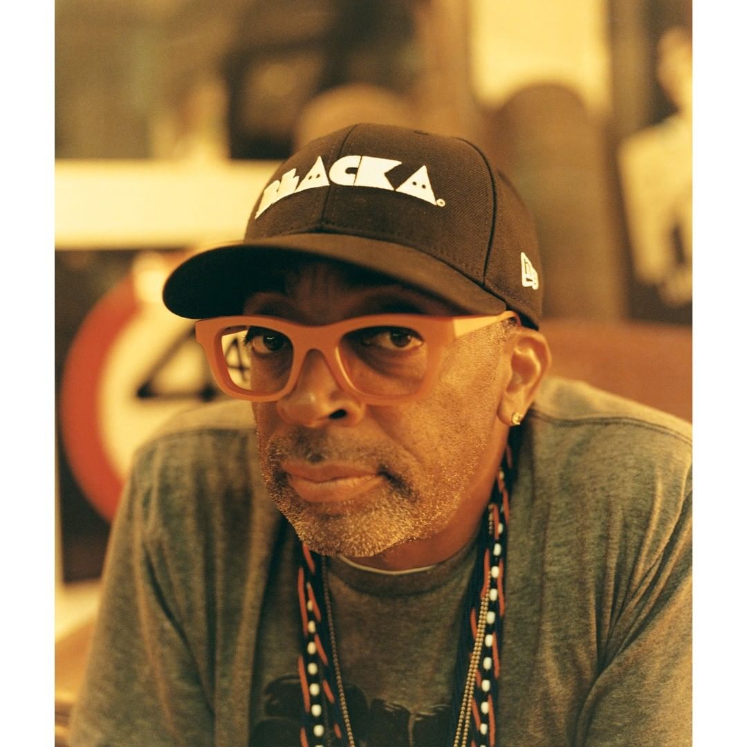 Portrait of Spike Lee wearing a baseball cap and orange glasses, gray t-shirt and beaded necklace, posing indoors with a neutral expression.