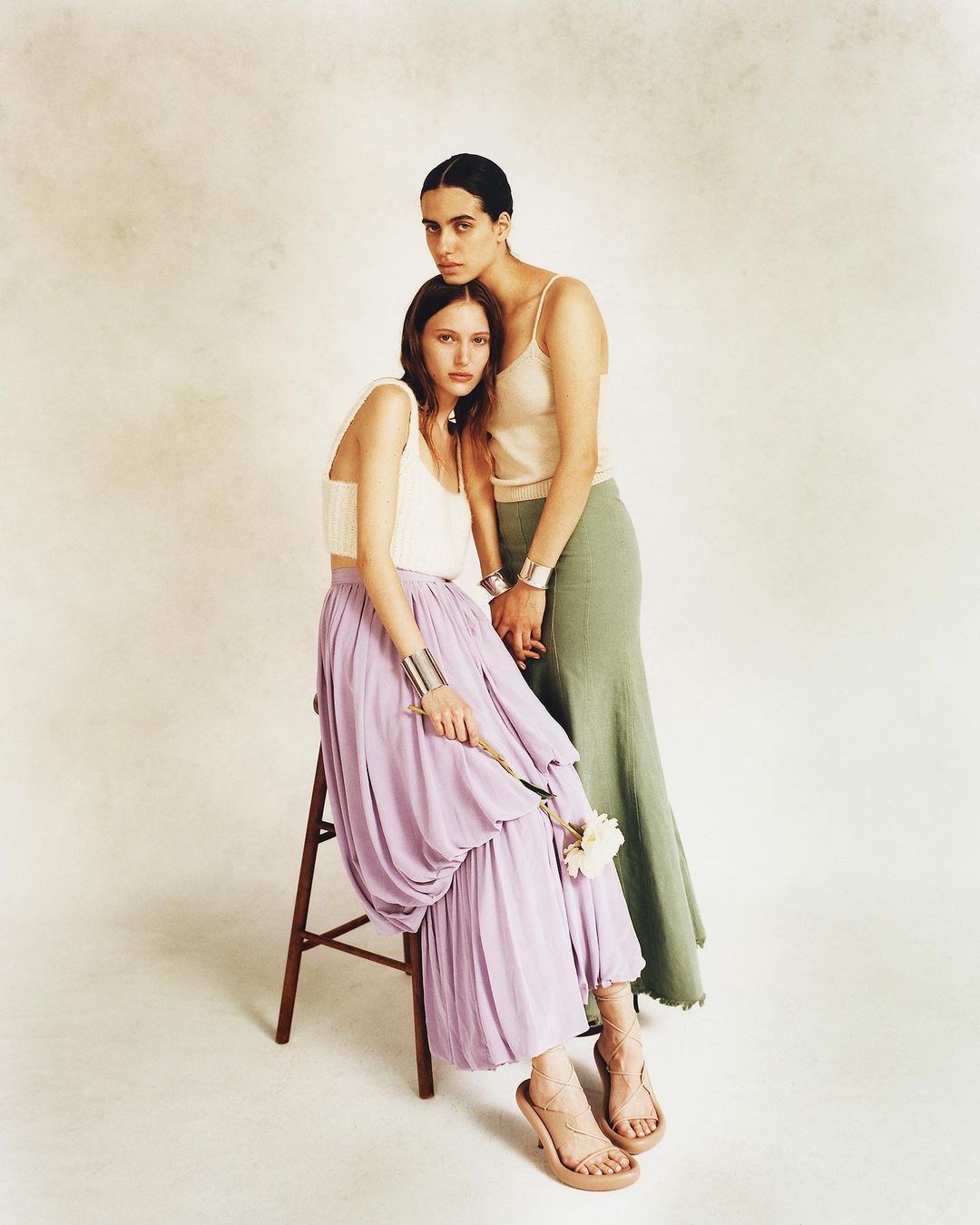 Two fashion models in elegant tops and flowing skirts, one seated on a stool holding a white flower, in a light studio setting.