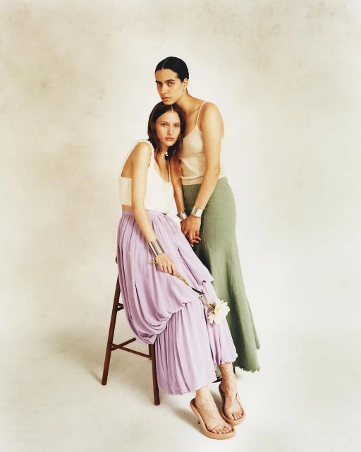 Two fashion models in elegant tops and flowing skirts, one seated on a stool holding a white flower, in a light studio setting.