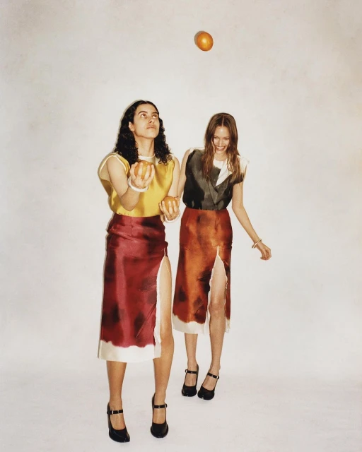 Two women in skirts and tops juggling oranges in a photo studio. One orange is in the air. Both women are smiling in this fashion editorial.