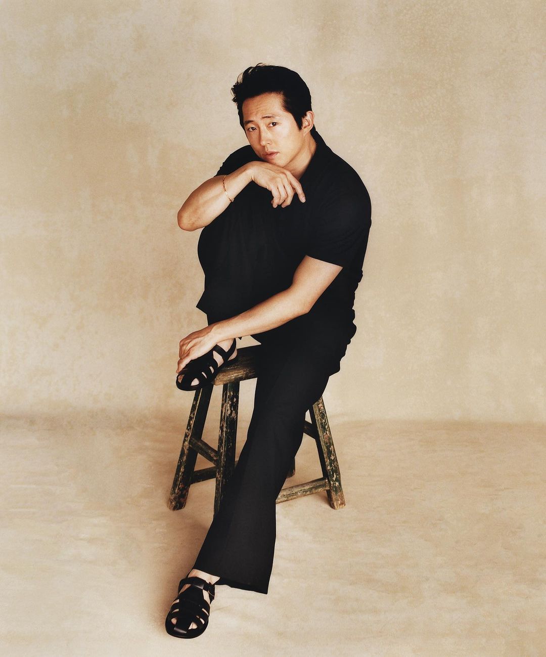 Portrait of an adult male sitting on a wooden stool in a photo studio, wearing black shirt, pants, and sandals against a neutral backdrop.