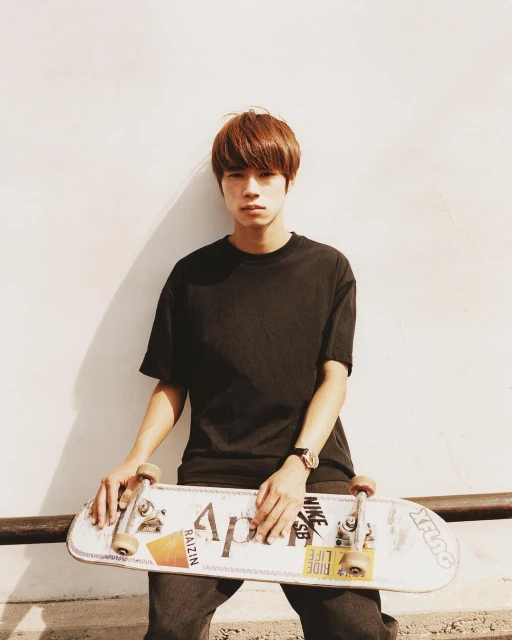 An Asian man in a black T-shirt holding a skateboard with logos on it, posing against a wall in sunlight, wearing a watch.