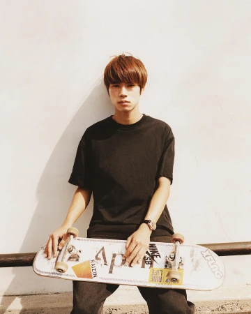 An Asian man in a black T-shirt holding a skateboard with logos on it, posing against a wall in sunlight, wearing a watch.