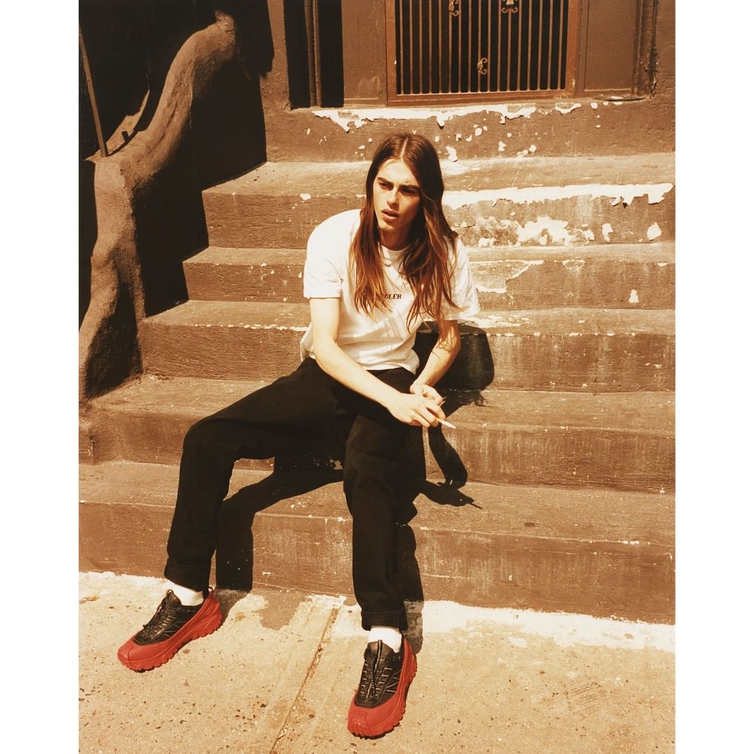 Young adult with long hair sits on concrete stairs in a casual white t-shirt, black pants, and bold red-soled shoes, holding a cigarette.