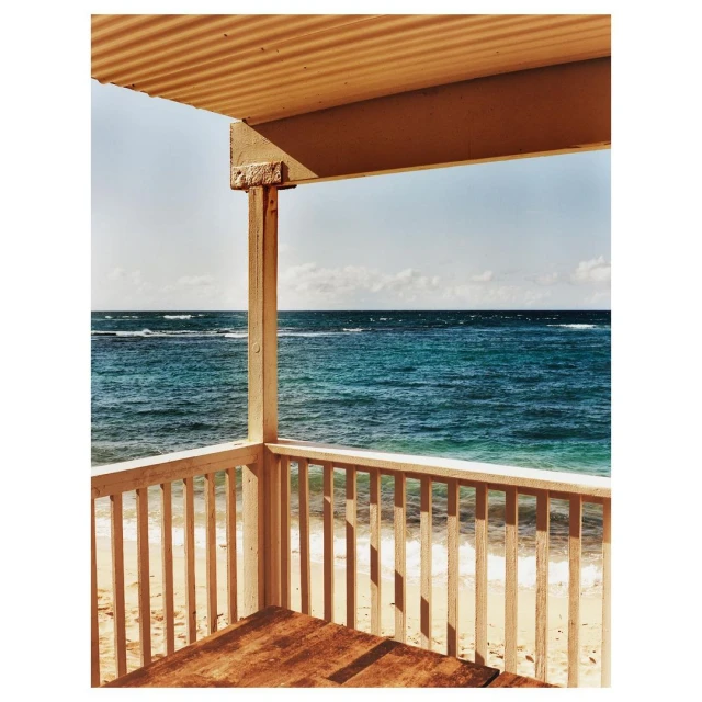 Seaside view from a covered porch, showing turquoise ocean water under a clear blue sky, with white sand visible in the foreground.