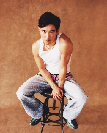 East Asian man in white tank top and patterned denim pants seated on a metal stool against a mottled brown backdrop