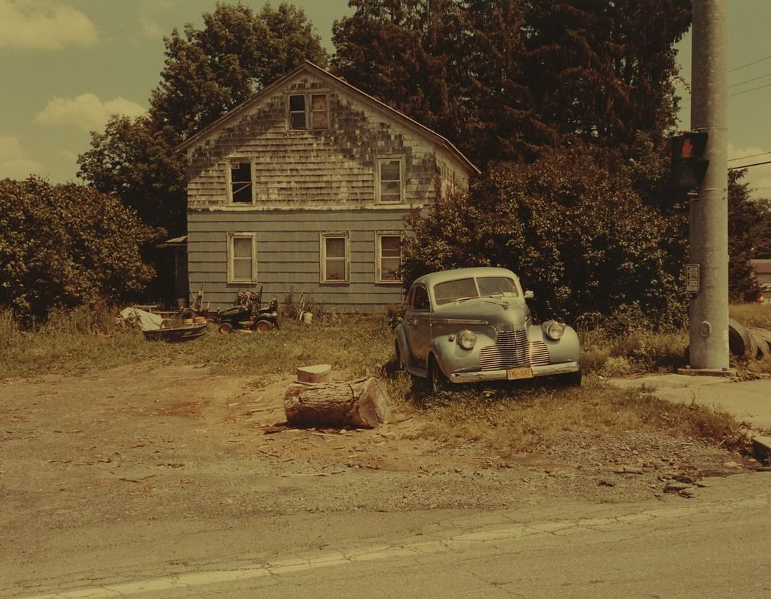 Vintage teal car parked on a grassy patch with a weathered two-story house in the background. Trees and sky visible.